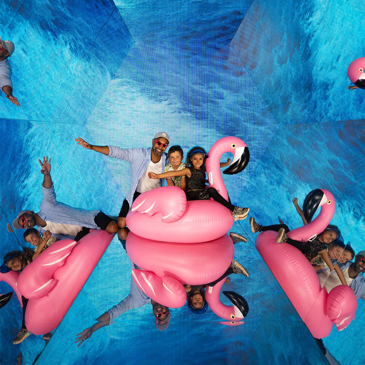 Two children seem to be floating on an inflatable pink flamingo while their father stands behind them in the middle of the Walk in Kaleidoscope. The screens that surround them emit a video of moving water.